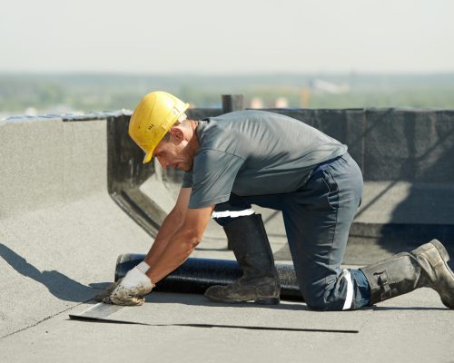 Flat roof covering works with roofing felt Roofer preparing part of bitumen roofing felt roll for melting by gas heater torch flame